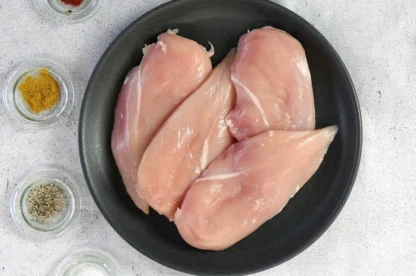 Three raw chicken breasts on a black plate, next to small glass bowls containing various spices and seasonings, on a light-colored surface.