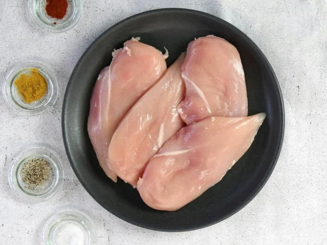 Three raw chicken breasts on a black plate, next to small glass bowls containing various spices and seasonings, on a light-colored surface.