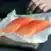 Two raw salmon fillets on parchment paper atop a wooden board, with carrots, a zucchini, and a person’s hand visible in the background.