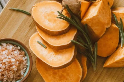 Sliced sweet potatoes on a wooden cutting board with sprigs of rosemary and a small bowl of coarse pink salt on the side.