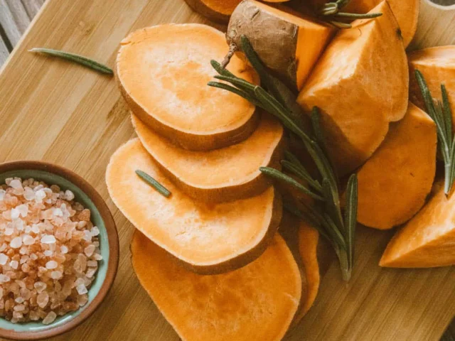 Sliced sweet potatoes on a wooden cutting board with sprigs of rosemary and a small bowl of coarse pink salt on the side.