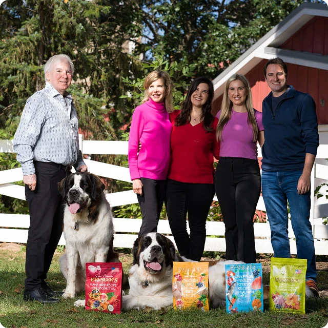 Five adults and two large dogs stand outside in front of a white fence and red barn, with five colorful bags of dog food displayed on the grass in front of them.