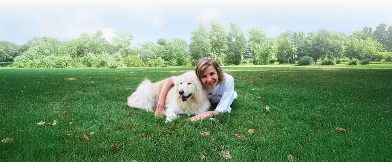 Woman lying on grass next to a fluffy white dog in a large, green park with trees in the background.