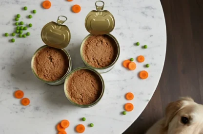 Two open cans of dog food are on a marble table, surrounded by scattered carrot slices and peas; a dog looks up from the floor beside the table.