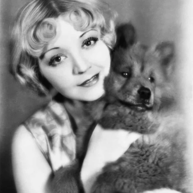 A woman with short, wavy hair holds a fluffy dog close to her face, both looking toward the camera. The photo is black and white and appears to be vintage.