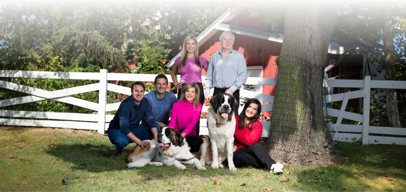 A group of six people and two large dogs pose together on grass in front of a white fence and a red barn, with trees in the background.