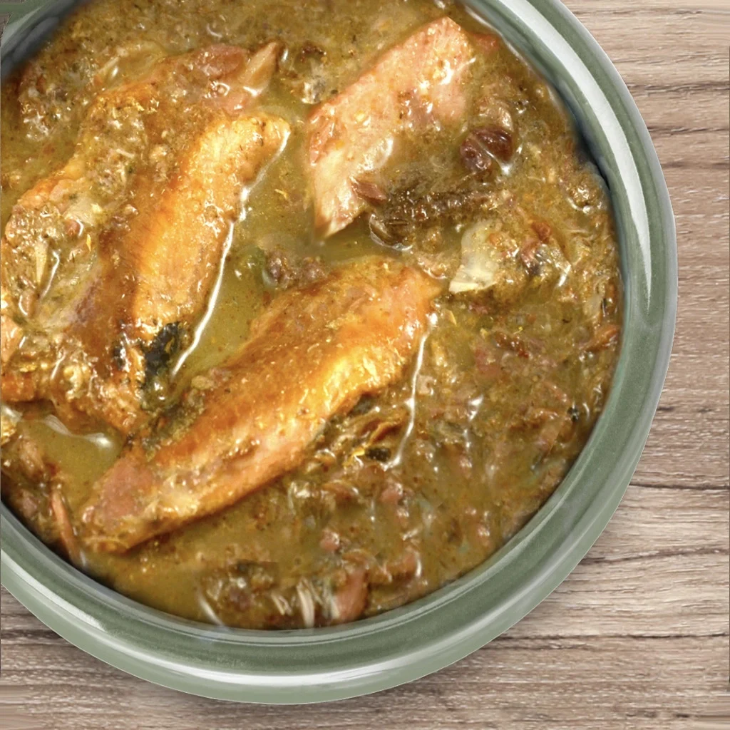 A bowl of soup with fish fillets and leafy greens in a light brown broth on a wooden surface.