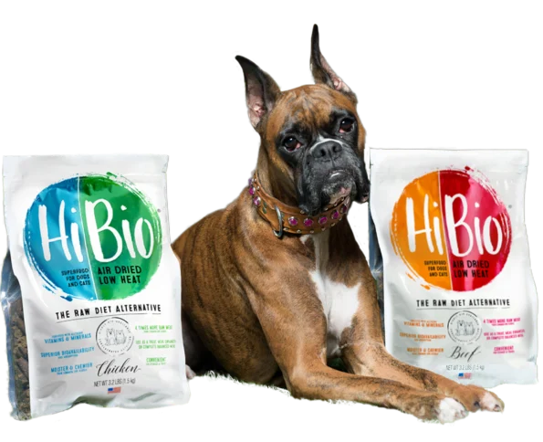 A brown and white dog lies between two bags of HiBio raw diet alternative dog food, one labeled Chicken and the other Beef, on a white background.